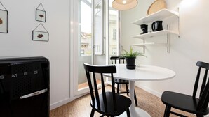 Una cocina moderna con armarios blancos, una mesa de comedor redonda, sillas negras, una lámpara colgante y una planta en maceta sobre la mesa.