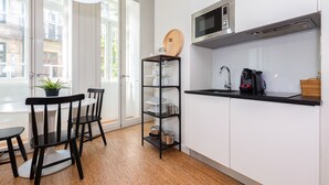 Una cocina moderna con armarios blancos, una encimera negra, microondas y cafetera. Hay una estantería de madera con vajilla y una planta en una mesa.