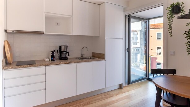 A modern kitchen with white cabinets, a coffee maker, and a sink. There is a wooden table and chairs in the dining area. - Feel Porto Downtown Theater Studio (Porto)