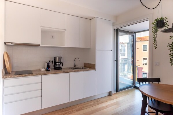 A modern kitchen with white cabinets, a coffee maker, and a sink. There is a wooden table and chairs in the dining area. - Feel Porto Downtown Theater Studio (Porto)