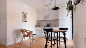 A modern kitchen with white cabinets, a wooden dining table, and black chairs. There is a hanging plant and a framed picture on the wall.