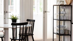 Een eetgedeelte met een tafel, stoelen, een bloempot en een metalen plank met servies en glazen.