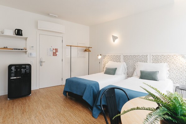 A hotel room with two beds, a desk, a chair, a small table, a potted plant, and a minibar.