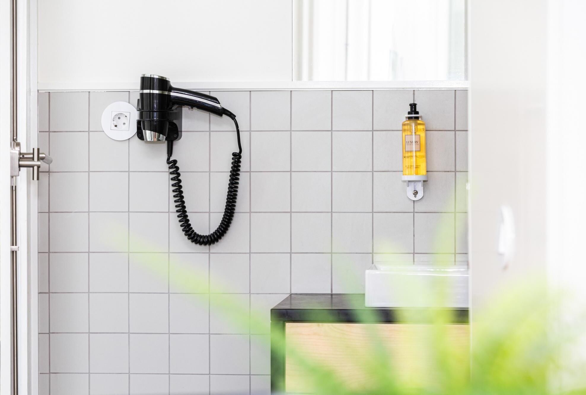 Een badkamer met een aan de muur bevestigde haardroger, een fles lotion en een betegelde wand.
