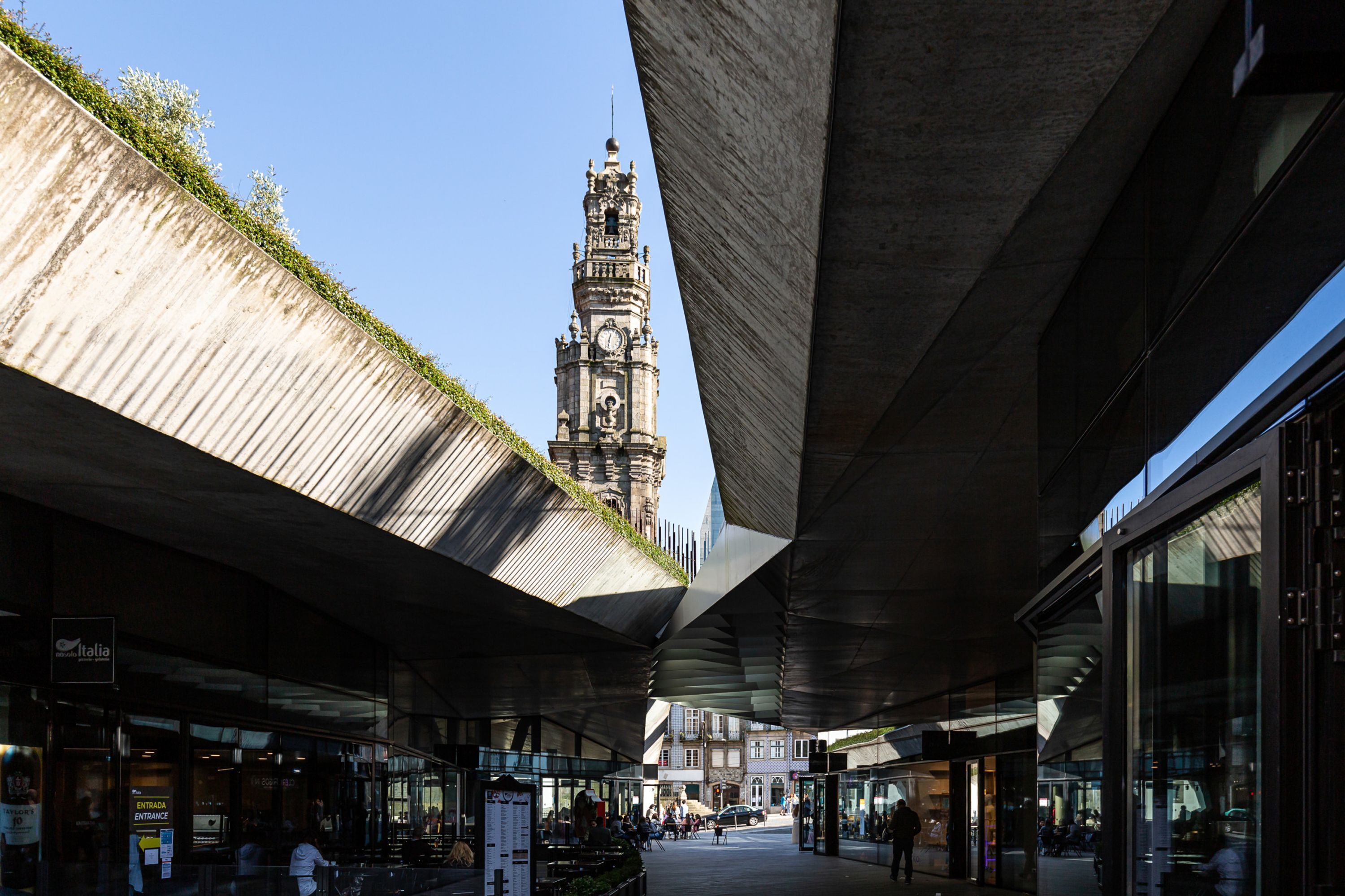 An urban pedestrian area with a historic clock tower in the background, modern architecture, and a covered walkway.