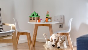 Una habitación con una mesa redonda de madera, dos sillas blancas, una maqueta de una casa, un juguete y una cesta con un peluche.