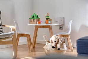 Quarto com uma mesa de madeira redonda, duas cadeiras brancas, uma maqueta de casa, um brinquedo e uma cesta com um bicho de pelúcia.