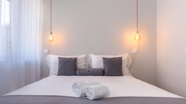 A neatly made bed with white linens and two rolled towels, two wall-mounted light fixtures, and a window with sheer curtains. - Feel Porto Downtown Harmony (Porto)