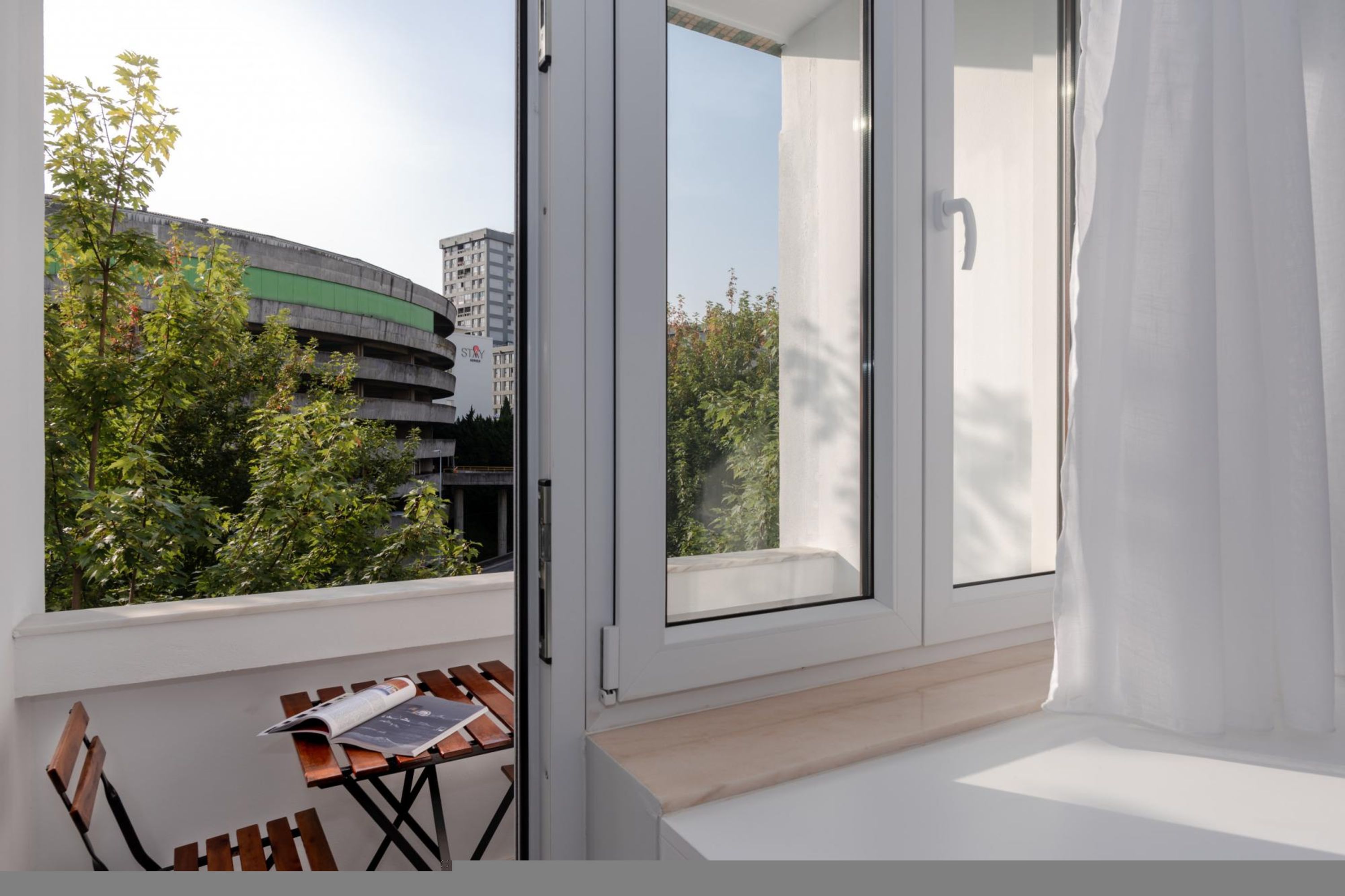 A balcony with a table and chairs, a window with curtains, and a view of a building and trees.