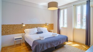 A modern bedroom with a large bed, a small bedside table, a wall-mounted light, and a window with curtains. - Feel Porto Downtown City Break (Porto)