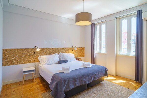 A modern bedroom with a large bed, a small bedside table, a wall-mounted light, and a window with curtains.