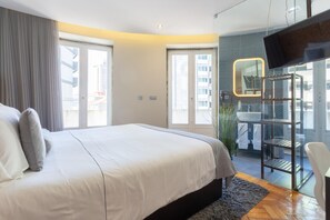 A modern hotel room with a large bed, a TV, a glass shower area, and a view of the city.
