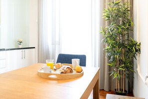 Un plateau de petit-déjeuner avec des croissants, un muffin, un verre de jus d’orange et une tasse, posé sur une table en bois dans une pièce agrémentée d’une grande plante et de rideaux.