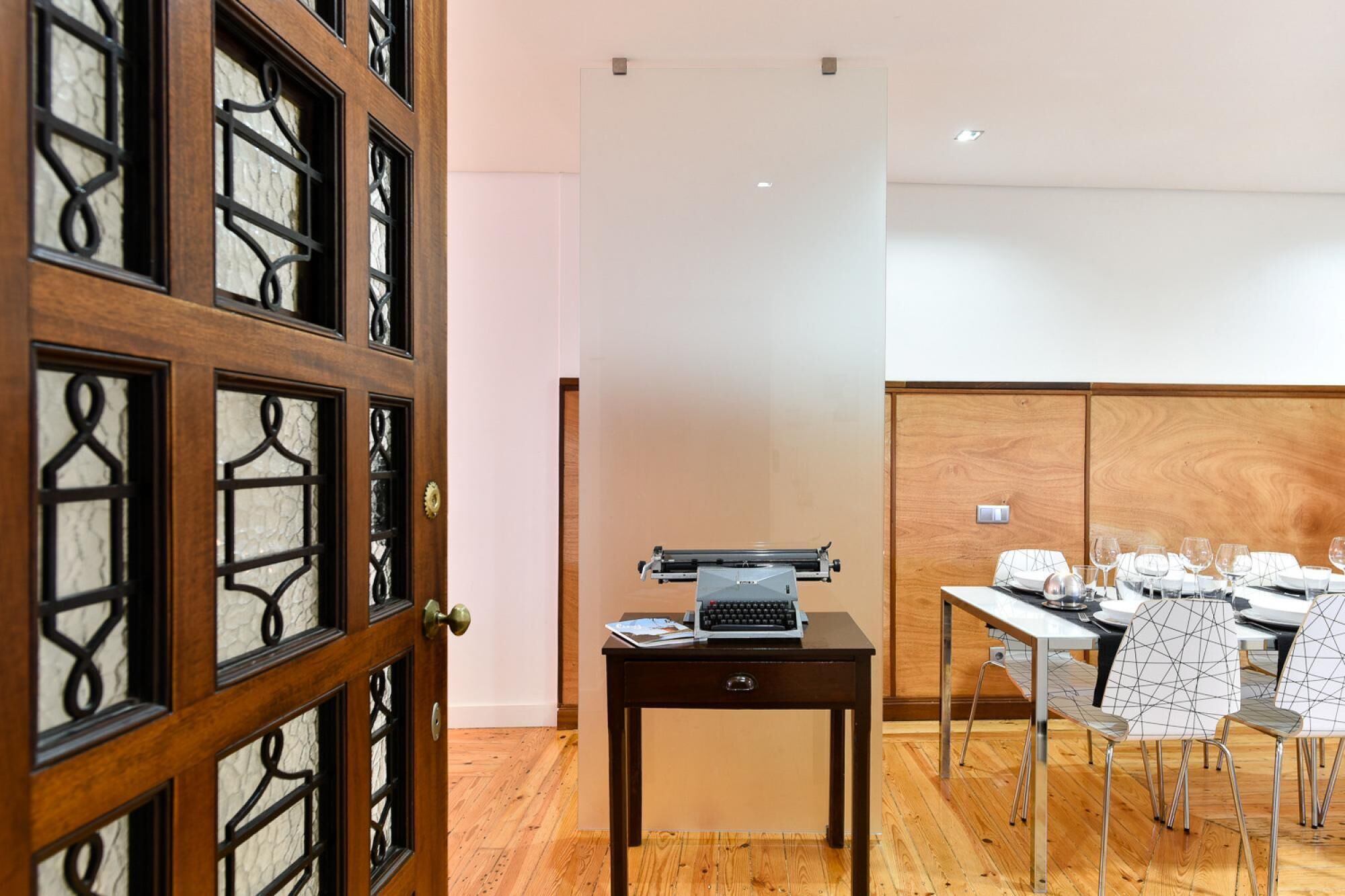 Une pièce avec une porte en bois, un bureau avec une machine à écrire et un coin repas avec des chaises et une table mise pour le repas.