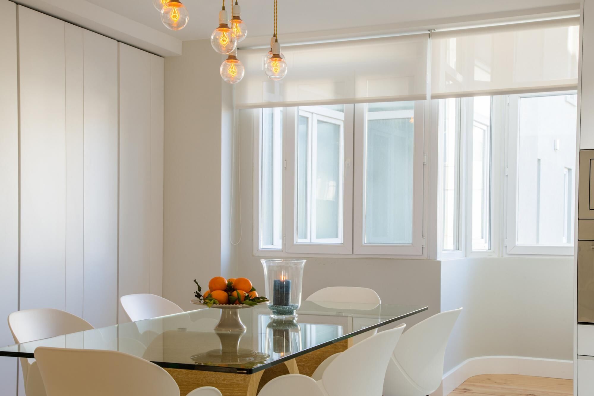 Un comedor moderno con una mesa de vidrio, sillas blancas y un florero con naranjas.