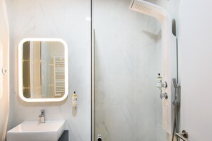 Een moderne badkamer met een wit wastafel, een spiegel met verlichting, een douche met handdouche en een zeepdispenser.