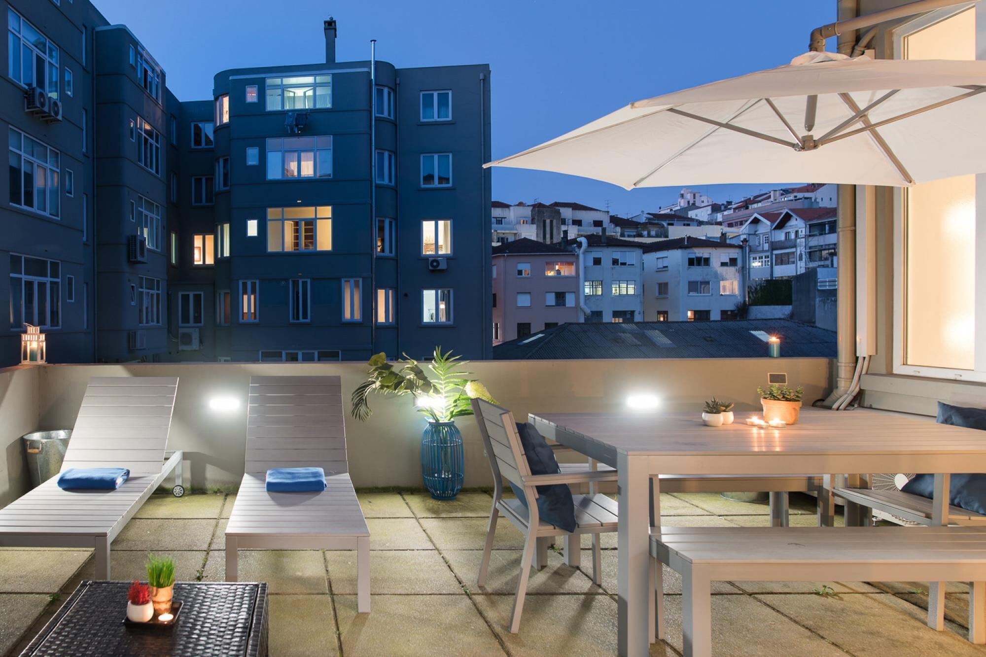 A rooftop terrace with a table, chairs, and a large umbrella. There are potted plants and a view of a cityscape in the background.