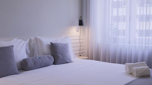 Ett hotellrum med en snyggt bäddad säng, en sänglampa och ett fönster med tunna, genomskinliga gardiner.