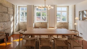 Une salle à manger avec une grande table en bois, six chaises, un lustre et de grandes fenêtres donnant sur un bâtiment au toit rouge.