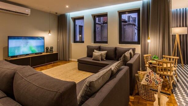 A modern living room with a large flat-screen TV, grey sofas, a wooden coffee table, and a view of buildings through the windows. - Feel Porto Vintage Townhouse (Porto)