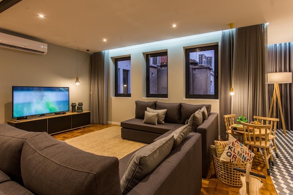 Una sala de estar moderna con un televisor de pantalla plana grande, sofás grises, una mesa de centro de madera y vistas a edificios a través de las ventanas.