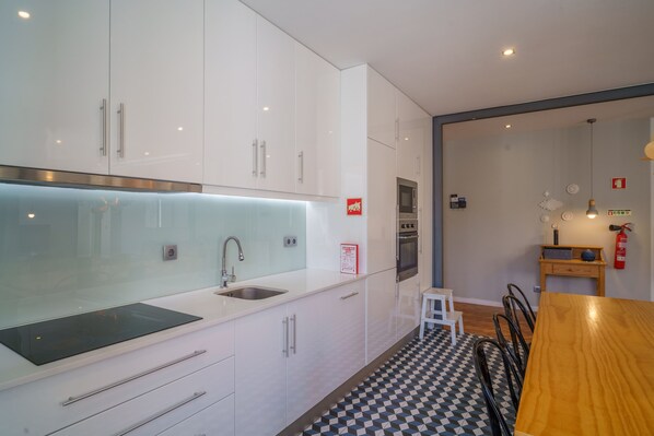 A modern kitchen with white cabinets, a stainless steel sink, and a built-in oven. There is a small dining area with a wooden table and chairs.