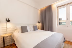 A hotel room with a large bed, two bedside lamps, a window with curtains, and a view of buildings outside.