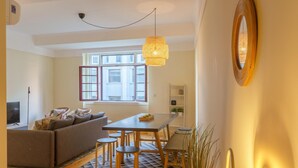 A modern living room with a sofa, dining table, and chairs. There is a TV, a window, and a hanging light fixture. - Feel Porto Downtown Woods Flat (Porto)