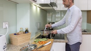 A modern kitchen with a person cooking, a wooden spice rack, a cutting board with vegetables, and a kettle. - Feel Corporate Housing Marqu s I (Porto)