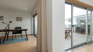 Una sala de estar moderna con piso de madera, un escritorio con una silla, y un amplio ventanal corredero que da a un área exterior con una mesa y sillas.