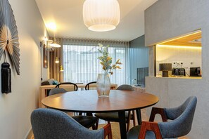 Un espace repas moderne avec une table ronde, des chaises, un vase de fleurs et une cuisine visible à travers une porte ouverte.