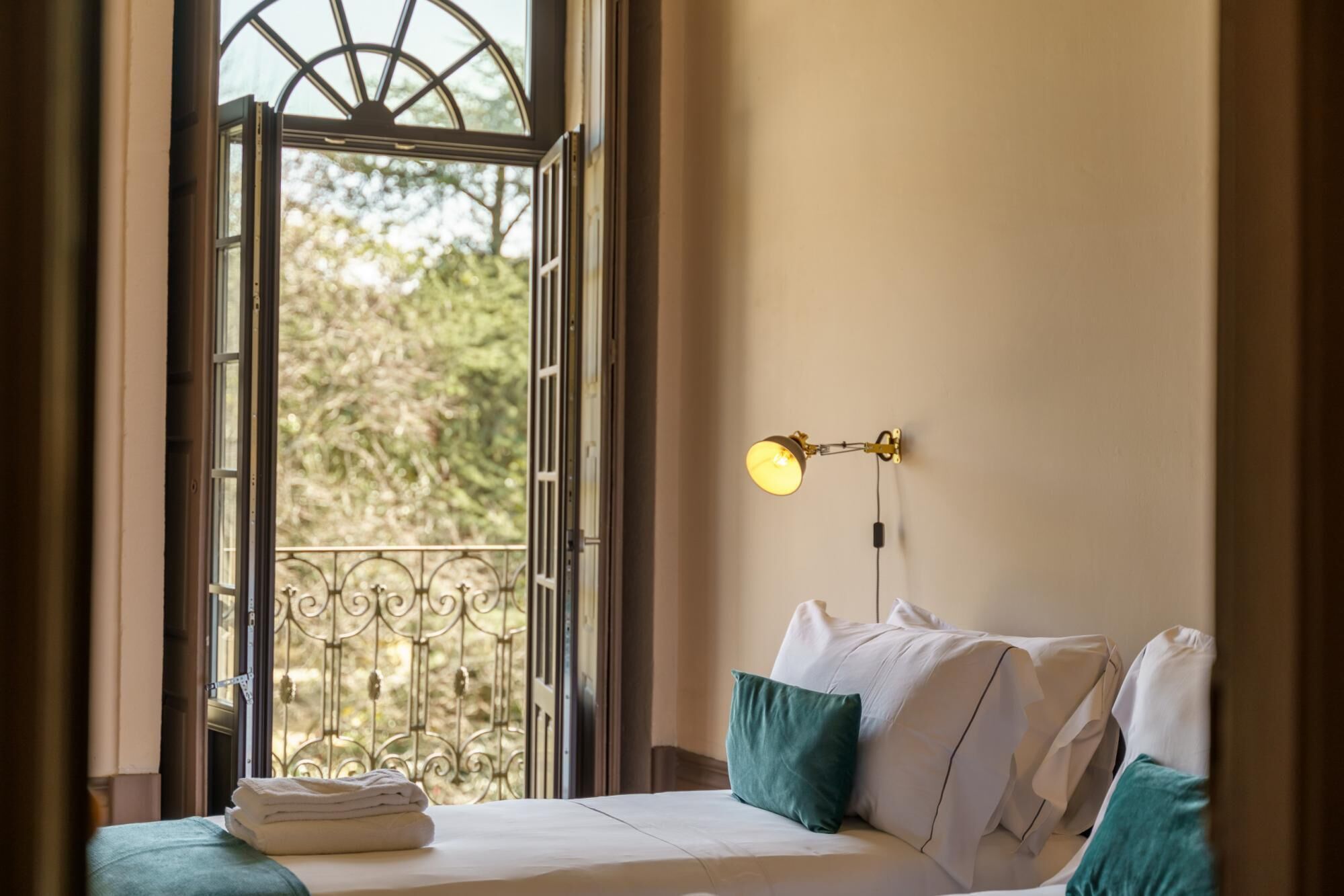 Una habitación de hotel con cama, ventana con vistas a árboles y lámpara fijada a la pared.