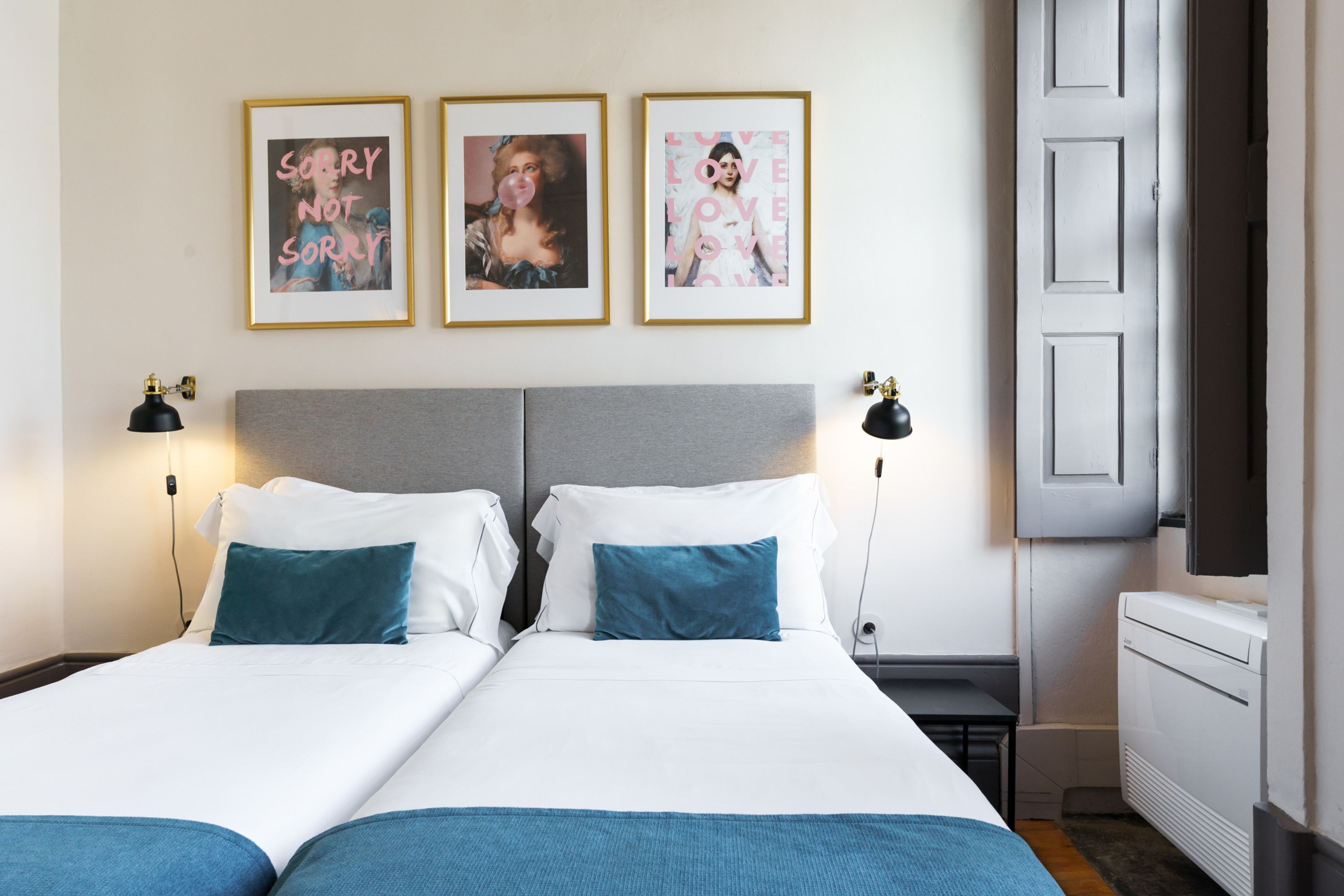 Habitación de hotel con dos camas, cabecero gris, ropa de cama blanca, almohadas turquesa y tres cuadros en la pared.