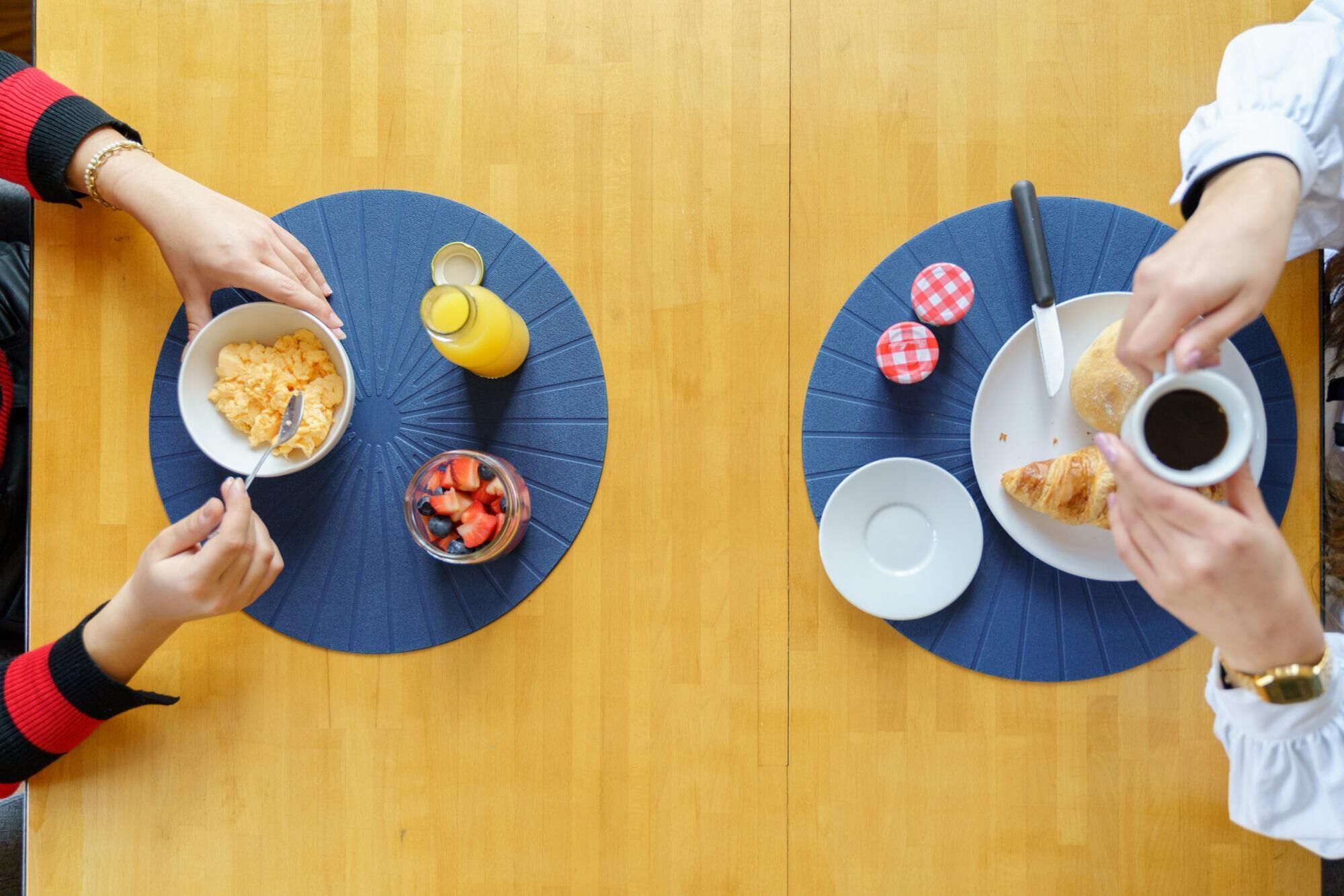 Una mesa de comedor con dos individuales azules, un tazón de huevos revueltos, un vaso de jugo de naranja, un frasco de bayas mixtas, un croissant y una taza de café.