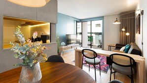 Uma sala de estar moderna com uma mesa de jantar de madeira, cadeiras, um sofá e uma televisão. Há uma janela grande que oferece vista para o exterior.