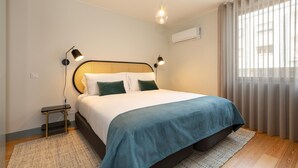 Quarto moderno com uma cama grande, luminárias de cabeceira, uma pequena mesa e uma janela com cortinas.