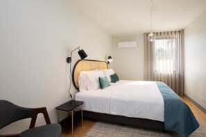 Un dormitorio ordenado con cama, mesitas de noche, una silla y una ventana con cortinas.