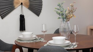 Ein Esstisch mit weißen Tellern, Schüsseln und Besteck, einer Vase mit getrockneten Blumen sowie zwei Weingläsern.