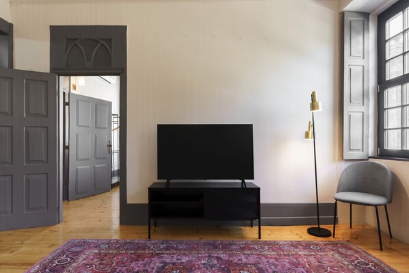 A modern living room with a flat-screen TV on a black stand, a patterned rug, a tall floor lamp, and a grey chair. - Feel Porto Antique Poveiros Flat I (Porto)