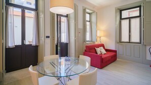 A modern living room with a glass table, white chairs, a red sofa, and a large window with curtains. - Feel Porto Matosinhos Ocean Flat V (Matosinhos)