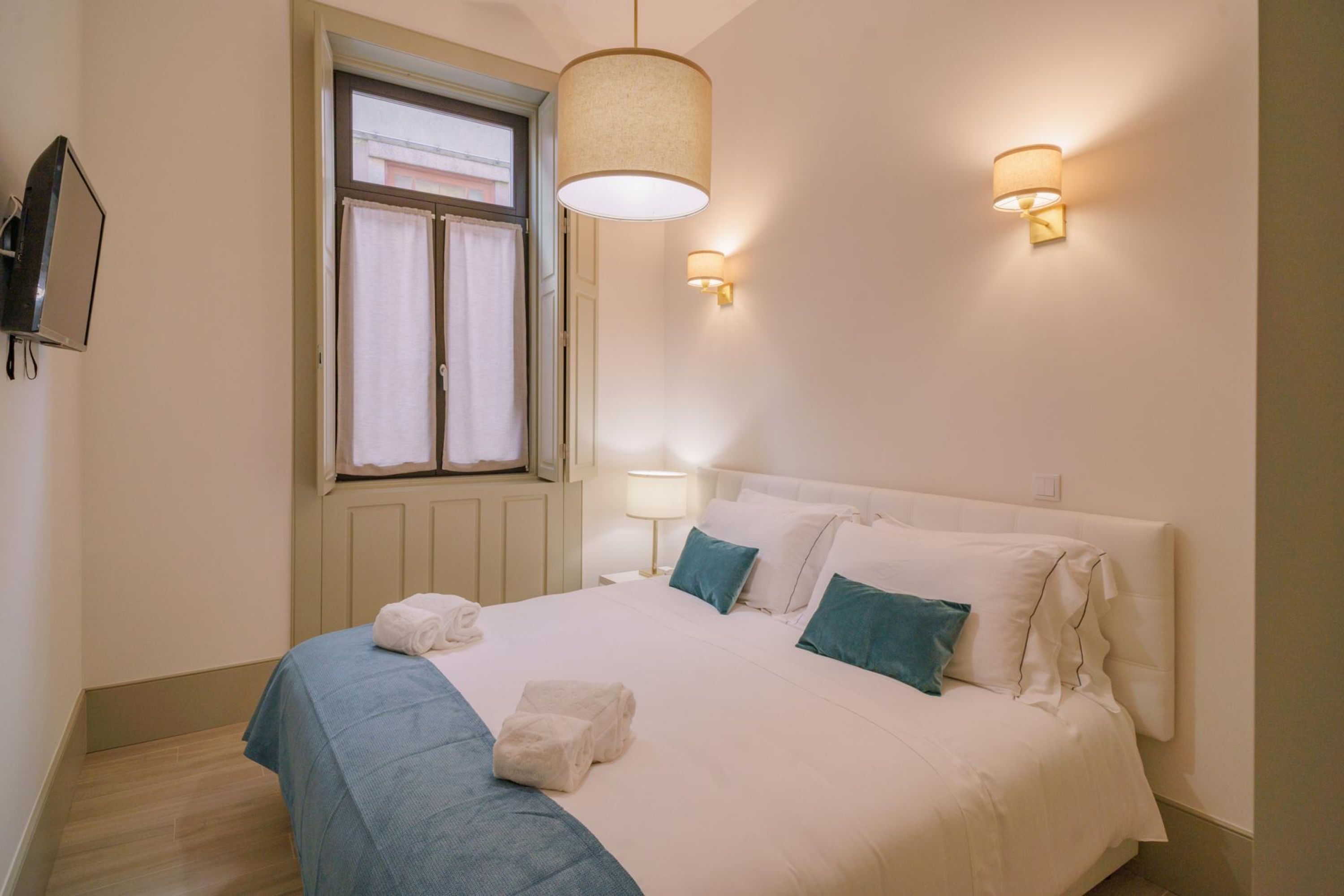 A bedroom with a bed, two wall-mounted lamps, a window with curtains, and a television on the wall.