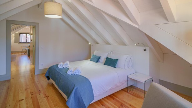 Un dormitorio con techo inclinado, una cama con colcha azul, almohadas blancas y una mesita de noche.