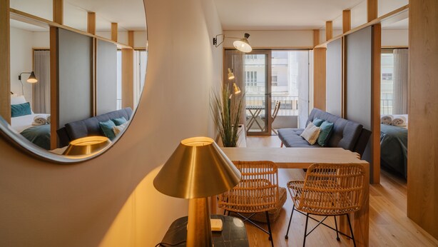 Una habitación de hotel moderna con un espejo redondo, una mesa de comedor con sillas, un sofá y un ventanal grande.