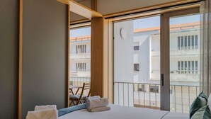 Habitación de hotel con cama, una silla y balcón con vistas a edificios.