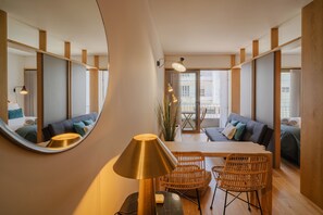 A modern hotel room with a round mirror, a dining table with chairs, a sofa, and a large window.