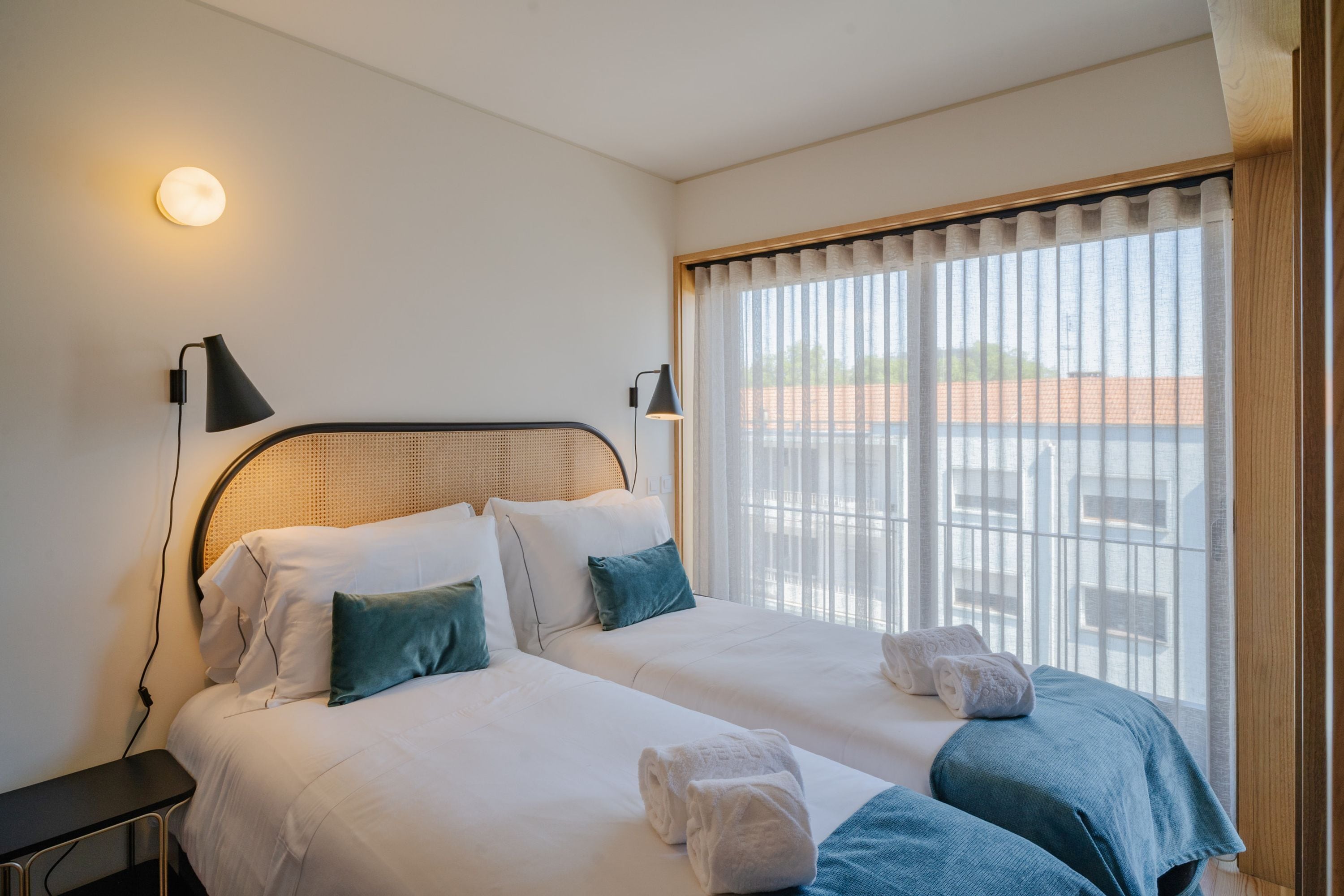 A hotel room with two beds, a large window with curtains, and a wall-mounted lamp.