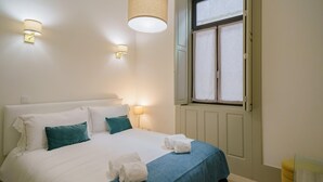 Una cama bien hecha con ropa de cama blanca y verde azulado, dos lámparas fijadas a la pared y una ventana con cortinas.