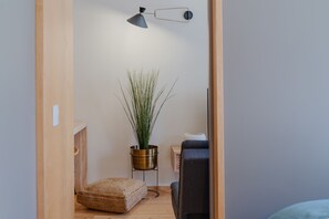 A small, cozy room with a wooden floor, a woven pouf, a potted plant, a desk with a chair, and a wall-mounted lamp.