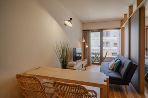 A modern living room with a wooden dining table, two wicker chairs, a grey sofa, and a large window offering a view of the city.