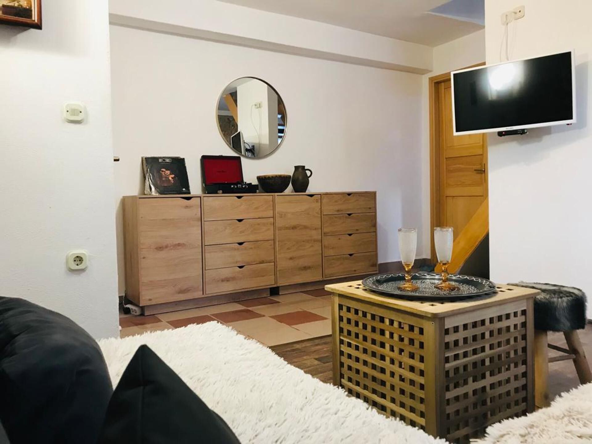 A living room with a wooden cabinet, a round mirror, a flat-screen TV, and a small table with glasses.